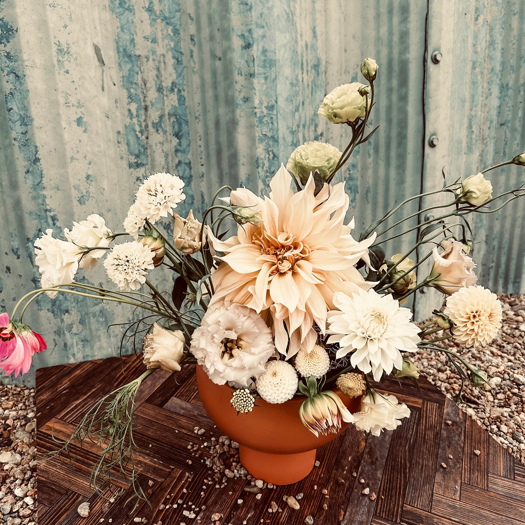 A bouquet of white and pale pink flowers in an orange vase on a wooden surface, set against a corrugated metal background.