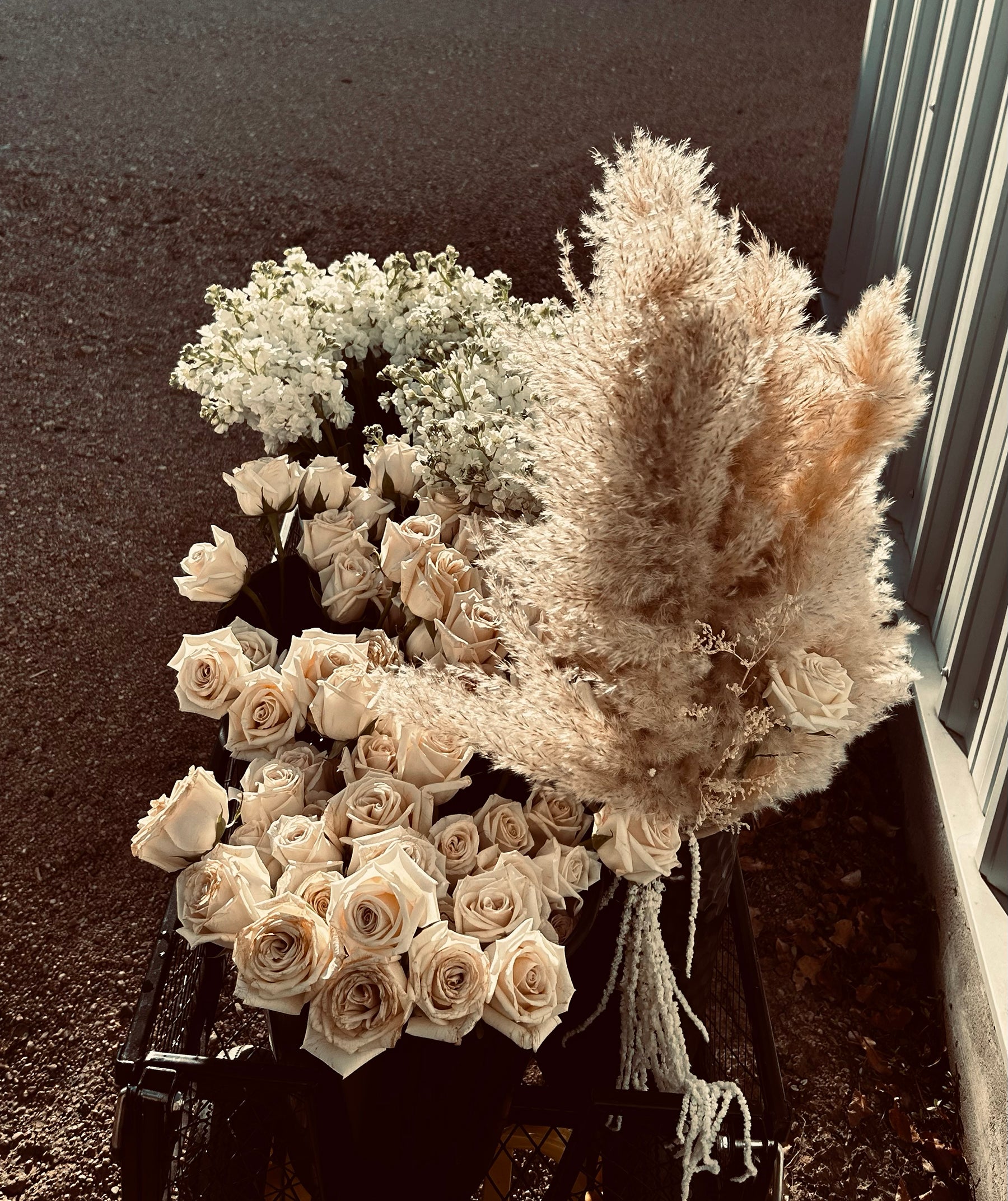 Bouquets of white roses, white flowers, and pampas grass arranged in black crates on a pavement, next to a building with corrugated metal siding.