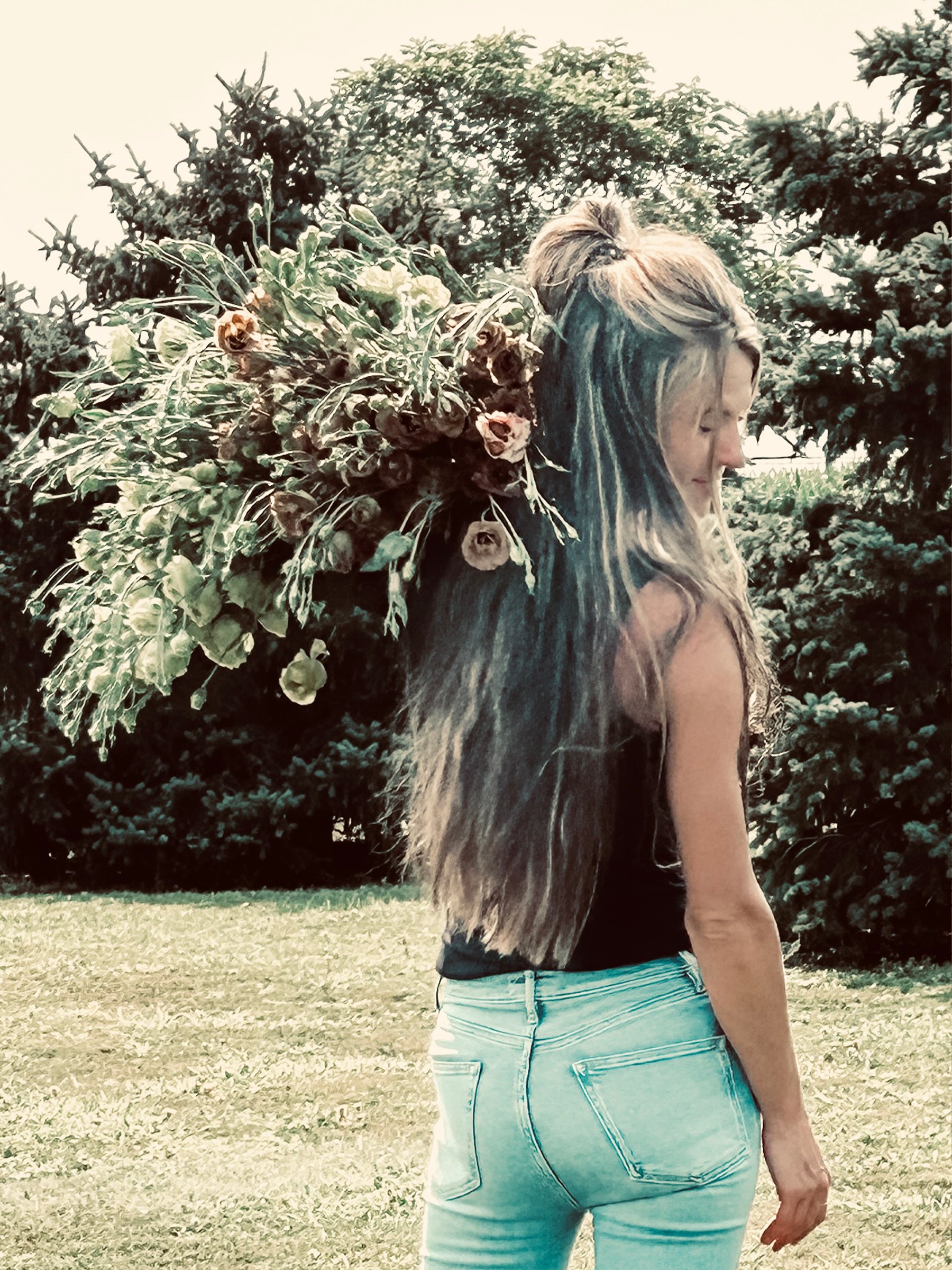 A woman with long hair carries a large bouquet of flowers over her shoulder while standing outdoors.