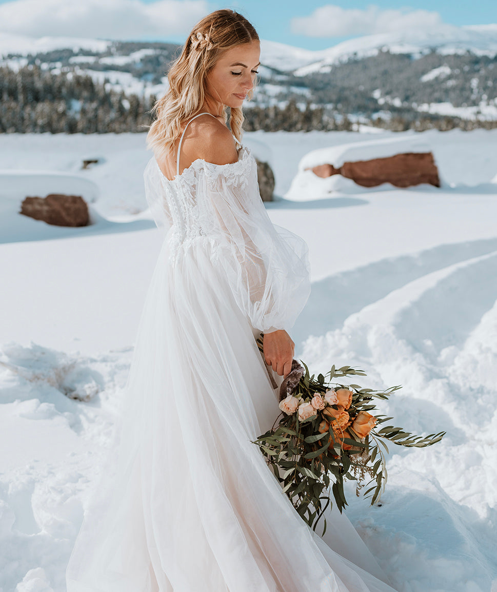 A bride in a white dress holds a bouquet of flowers, standing in a snowy landscape with mountains in the background.
