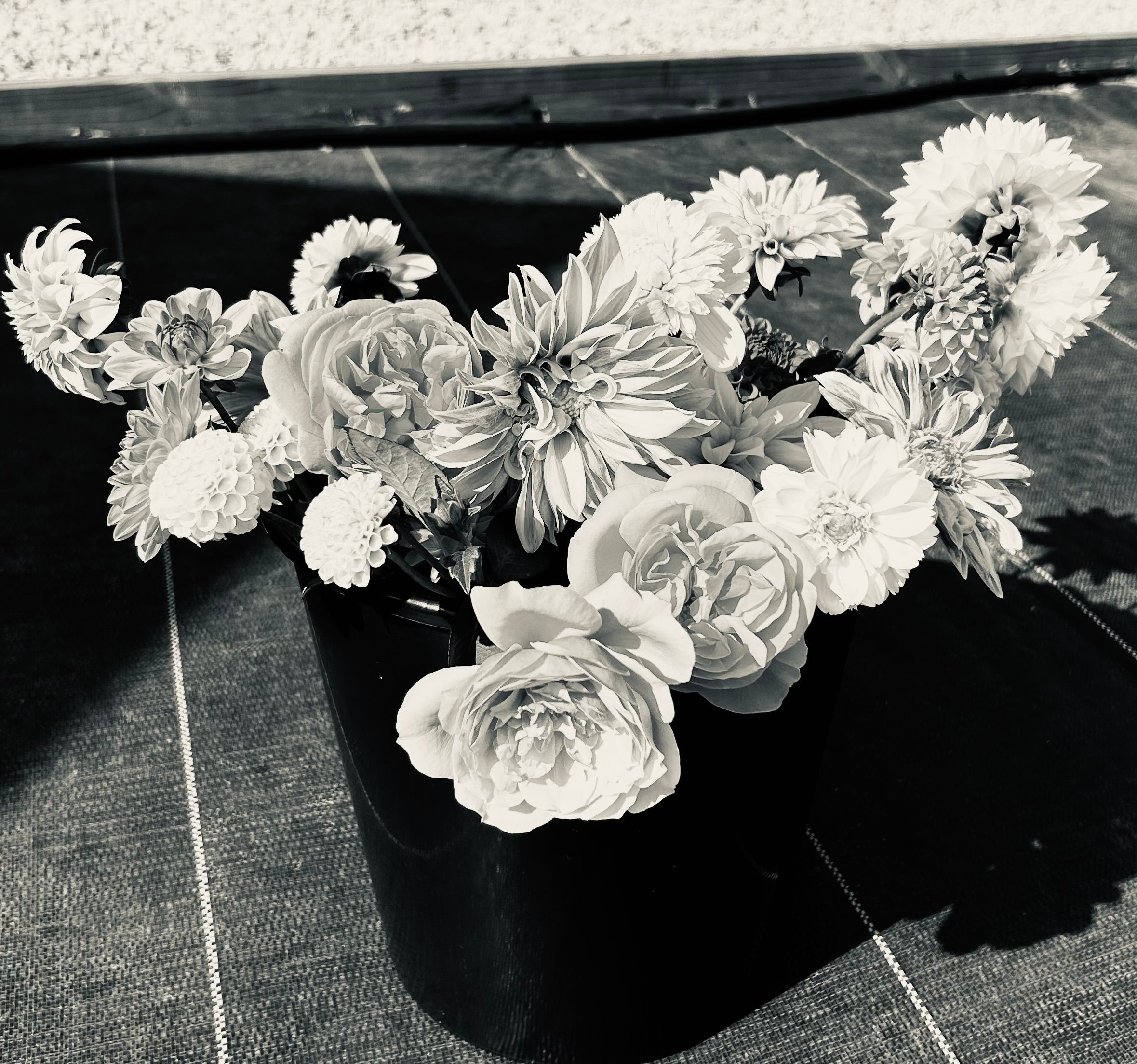 Black and white photo of a black vase filled with various flowers, including roses and dahlias, placed on a patterned surface.