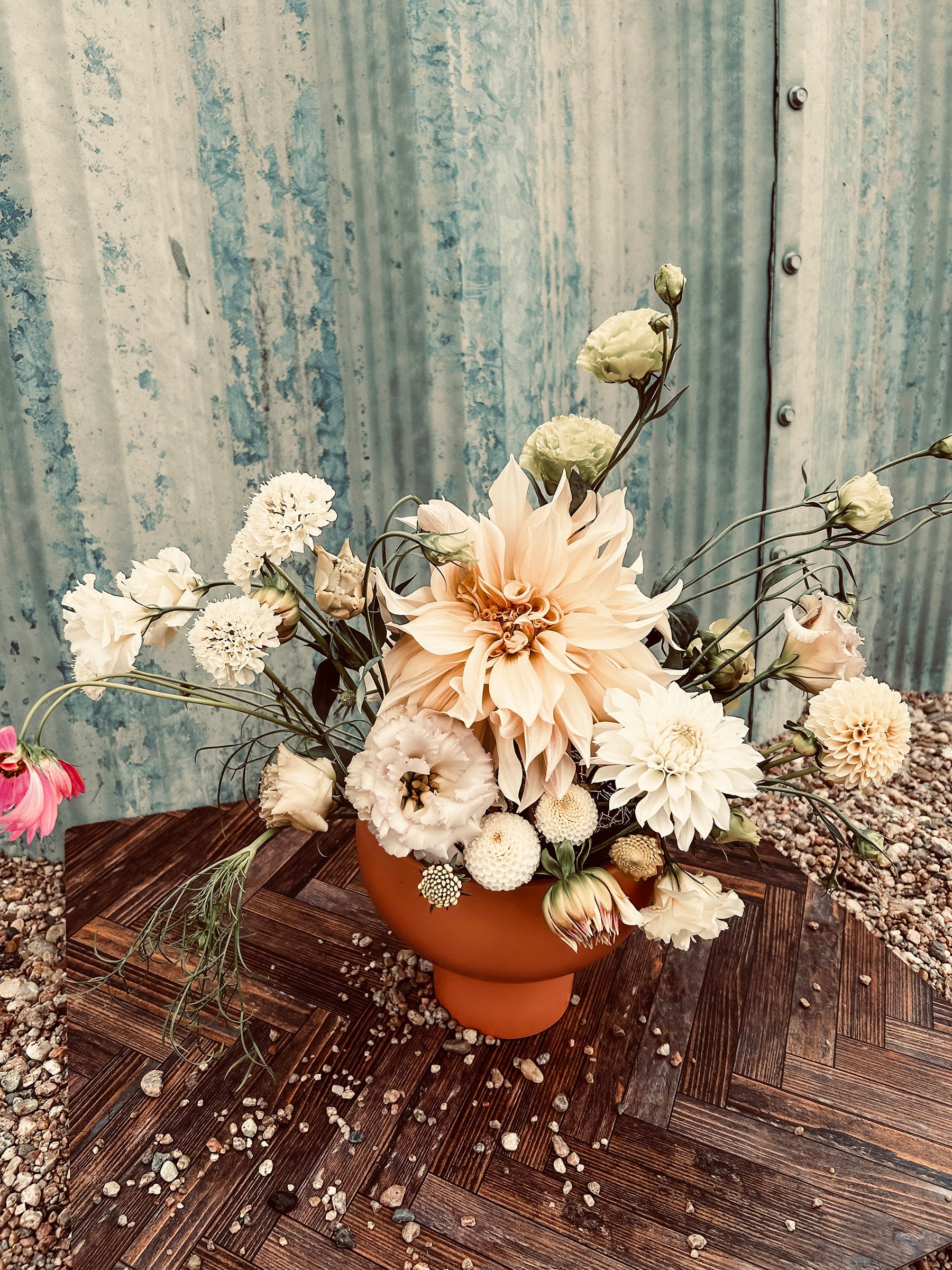 A bouquet of white and pale pink flowers in an orange vase on a wooden surface, set against a corrugated metal background.