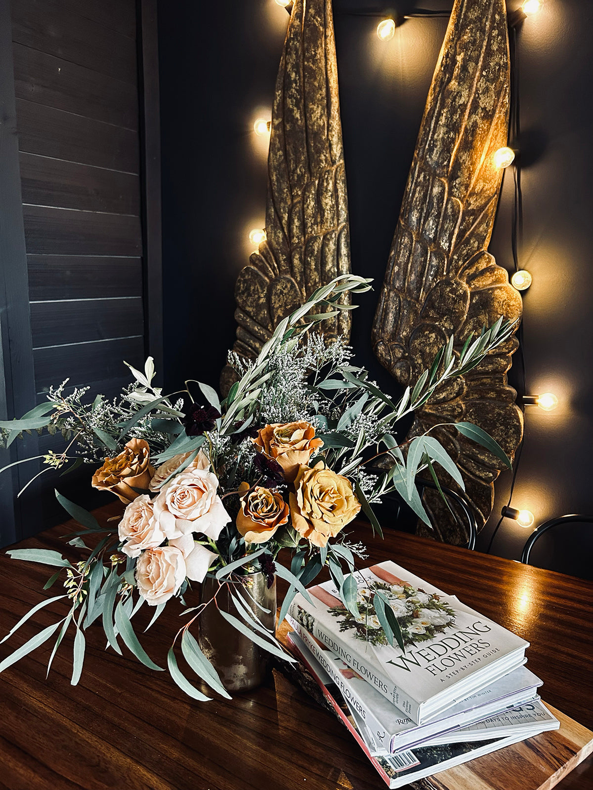 Floral arrangement with orange and white roses on a wooden table, next to books about weddings. Background features angel wing decor and string lights on a dark wall.