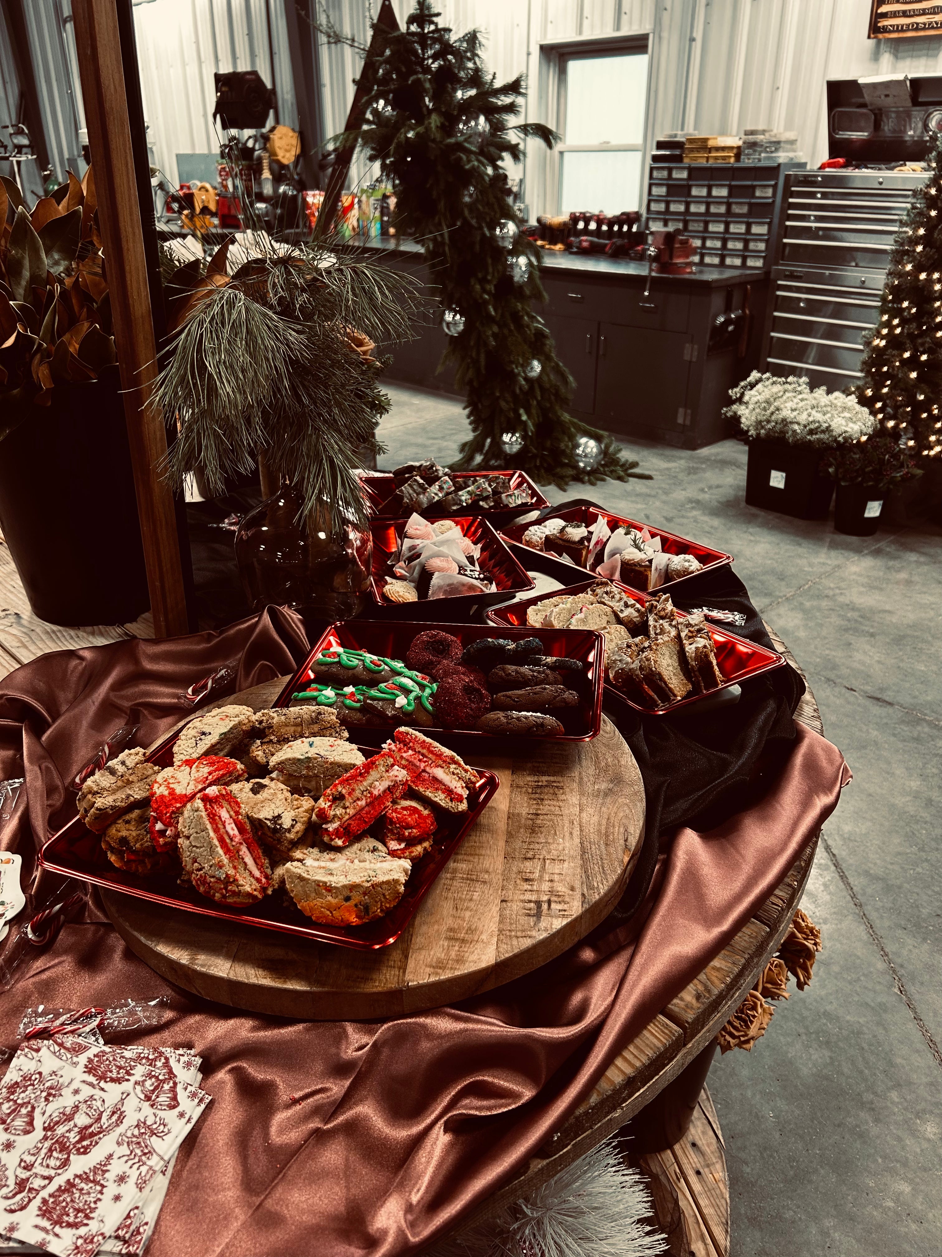 Assorted holiday cookies and treats on a decorated table with trees in the background inside a workshop.