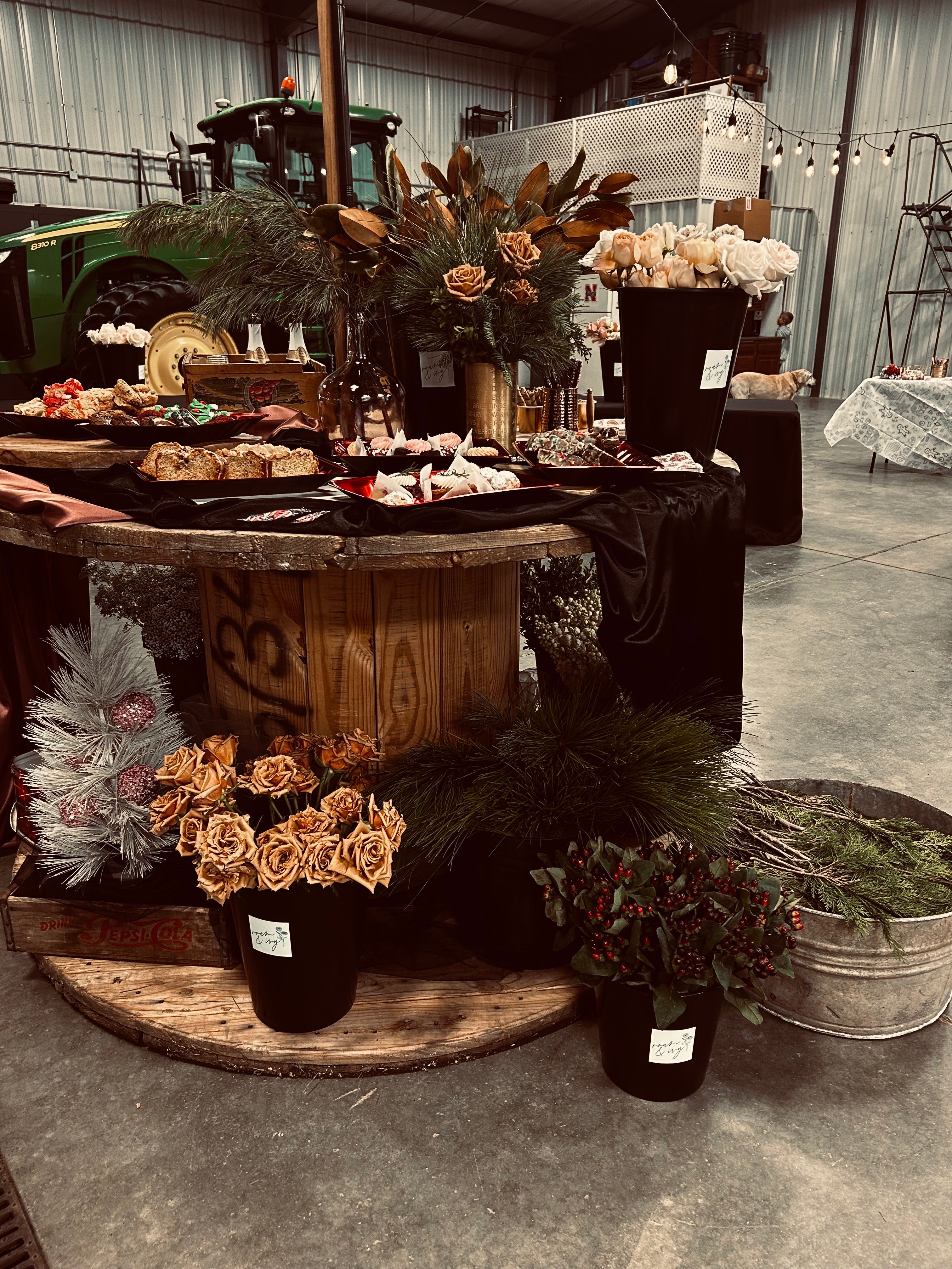 Display of flowers on a wooden table with black cloth, surrounded by greenery and floral arrangements in a warehouse setting.