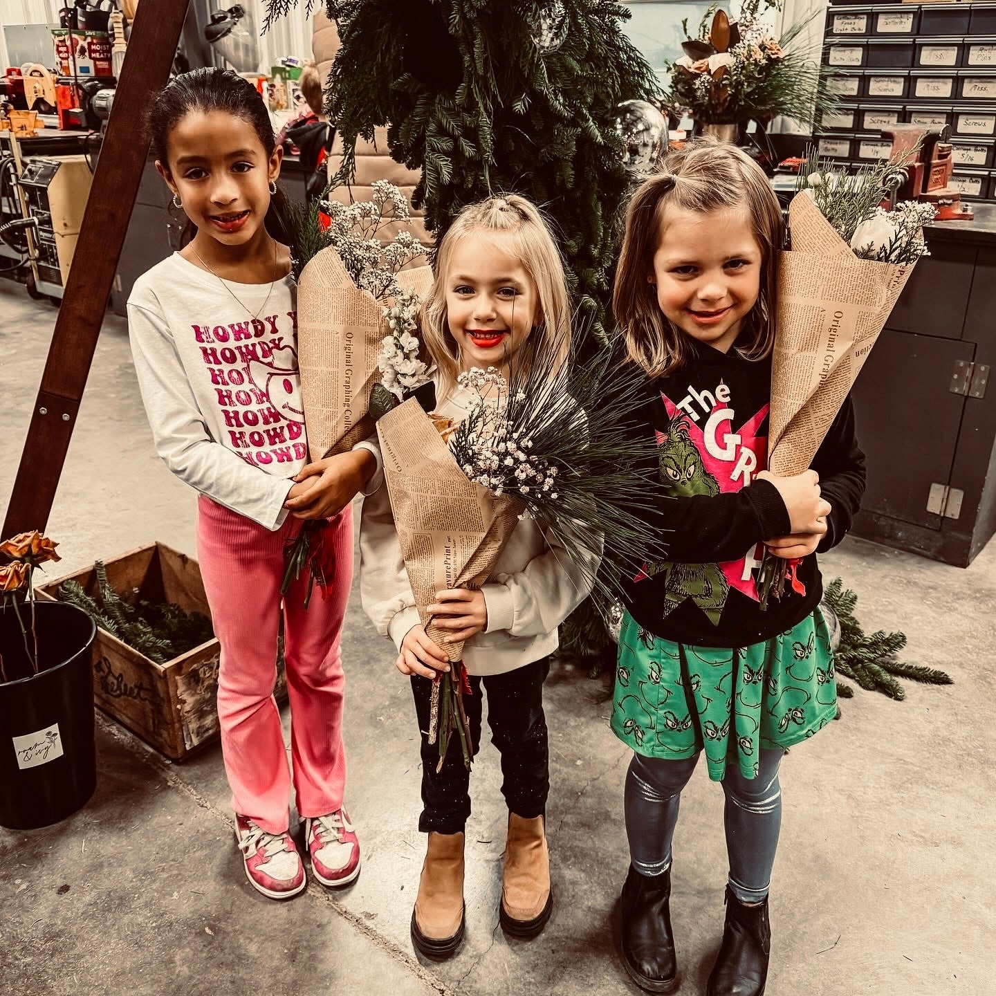 Three girls standing in a workshop, each holding a bouquet wrapped in brown paper, with tools and storage boxes in the background.