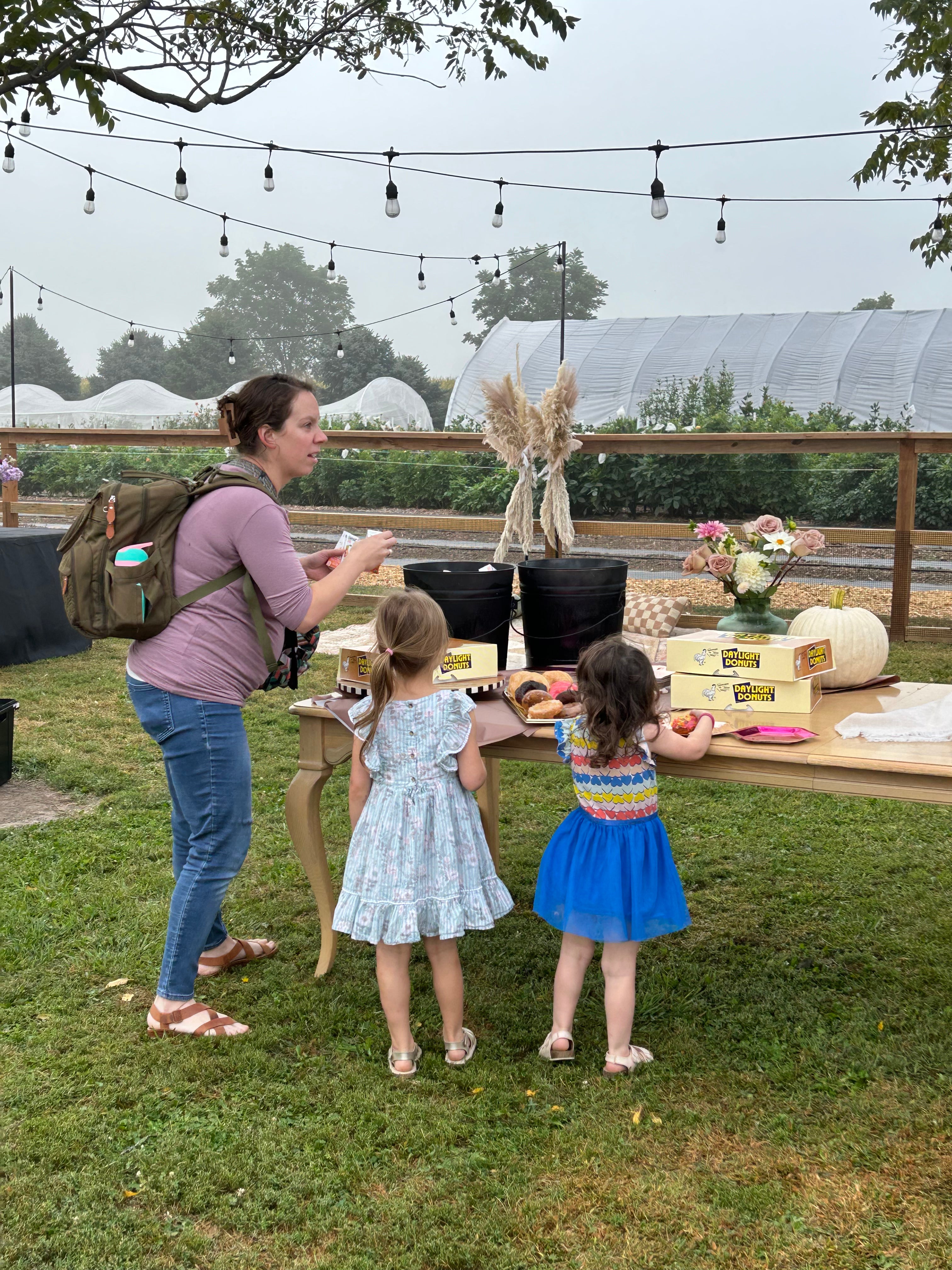 A woman with a backpack and two young girls stand by a table outdoors, eating snacks. The table has doughnut boxes, a floral arrangement, and a pumpkin. String lights hang above.
