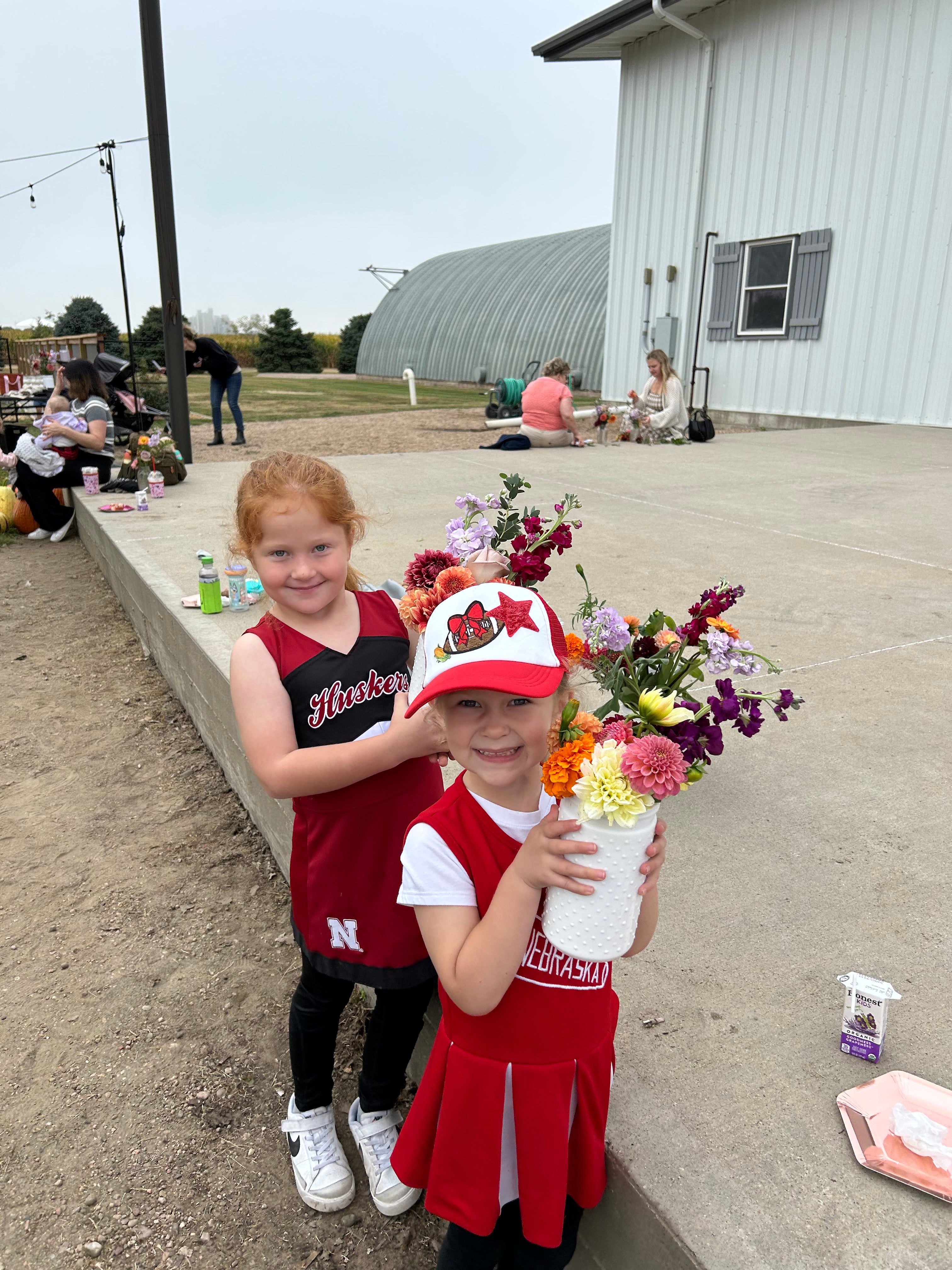 Two young girls in Nebraska cheer outfits, one holding a bouquet of flowers, standing outside near a white building. Other people are gathered in the background.