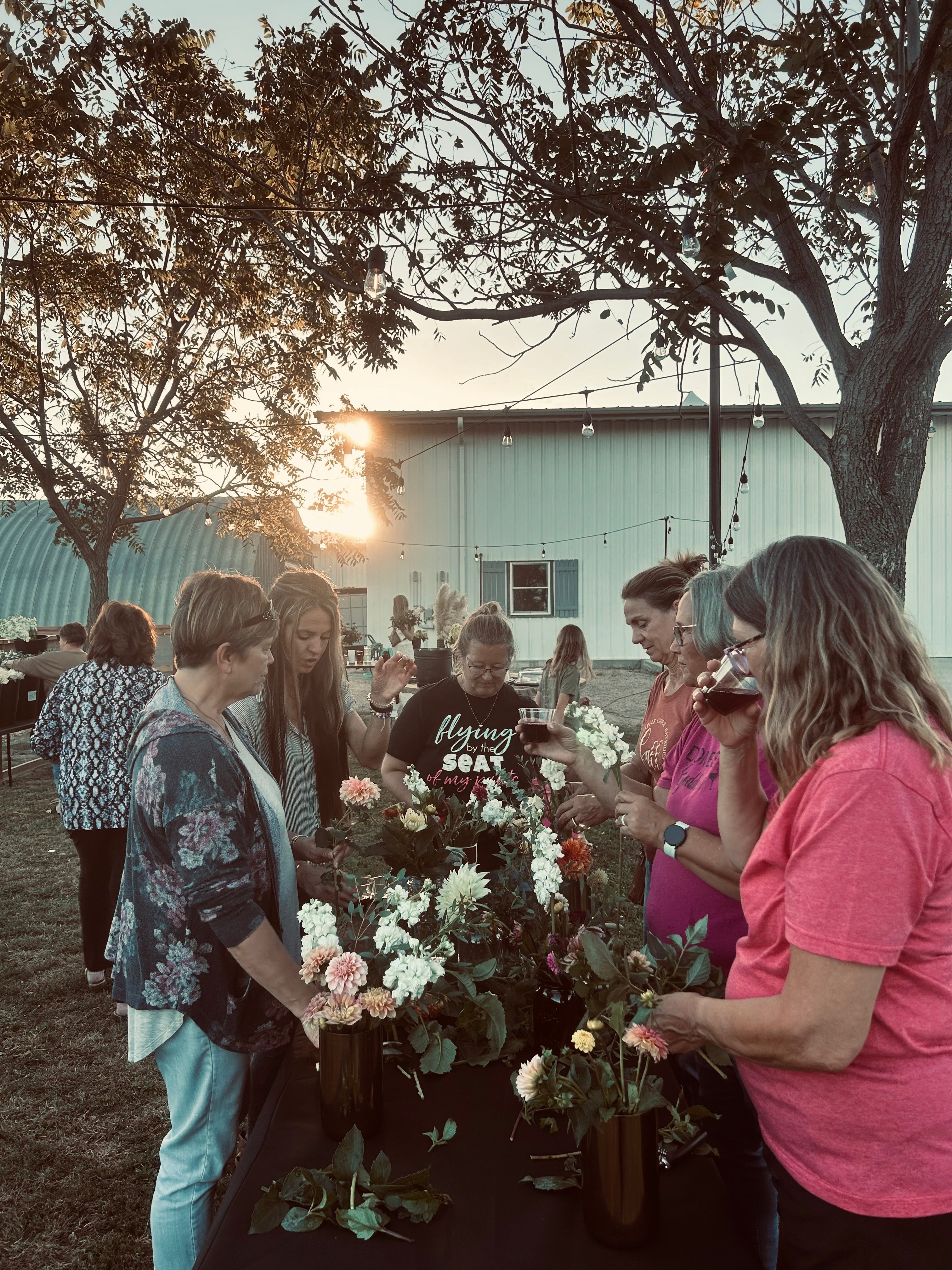 A group of people gather around a table arranging flowers outdoors near a building at sunset.