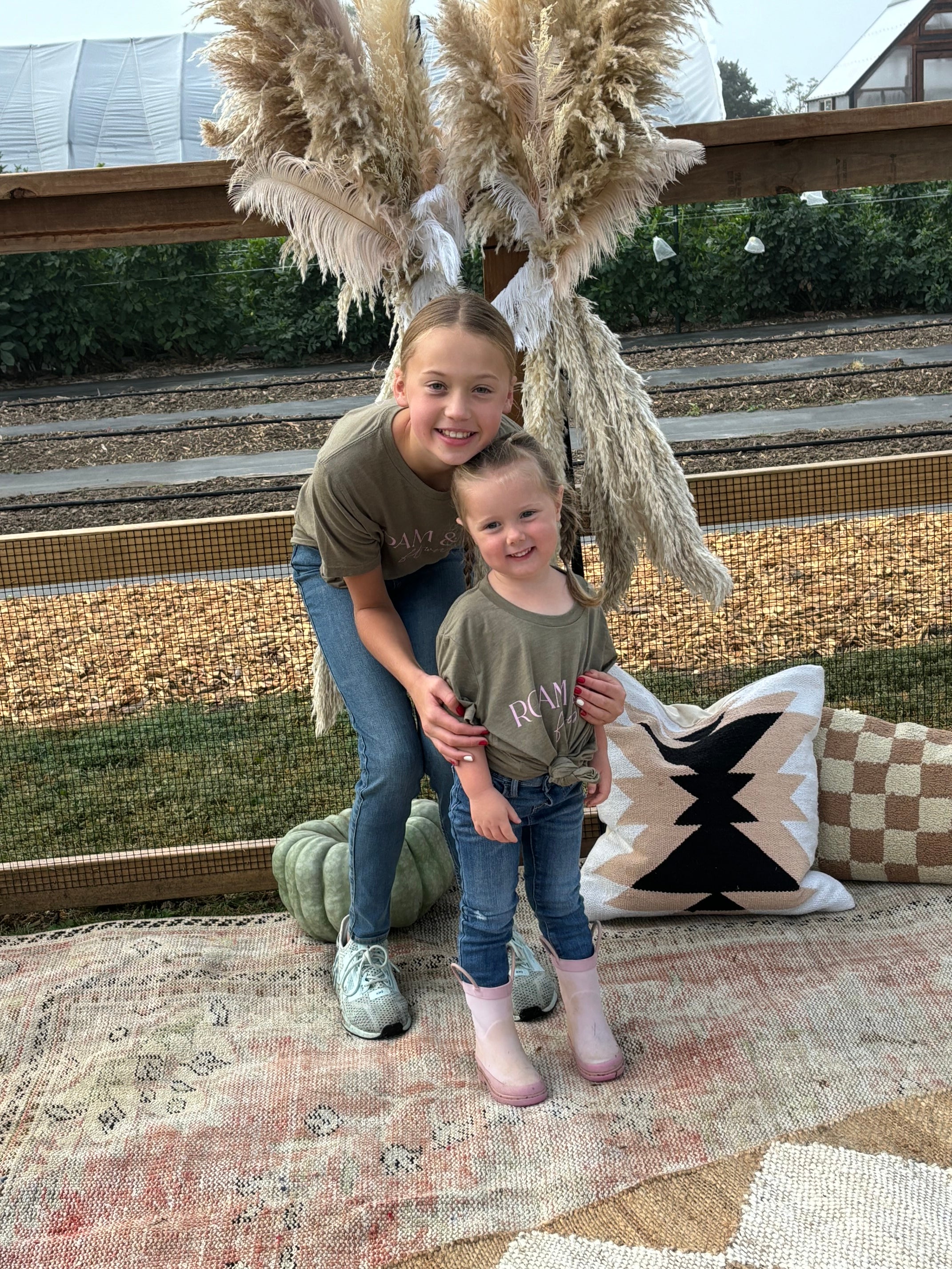 Two children smiling, standing on a patterned rug outdoors, with pampas grass decor and a rustic wood backdrop. The older child is hugging the younger from behind.