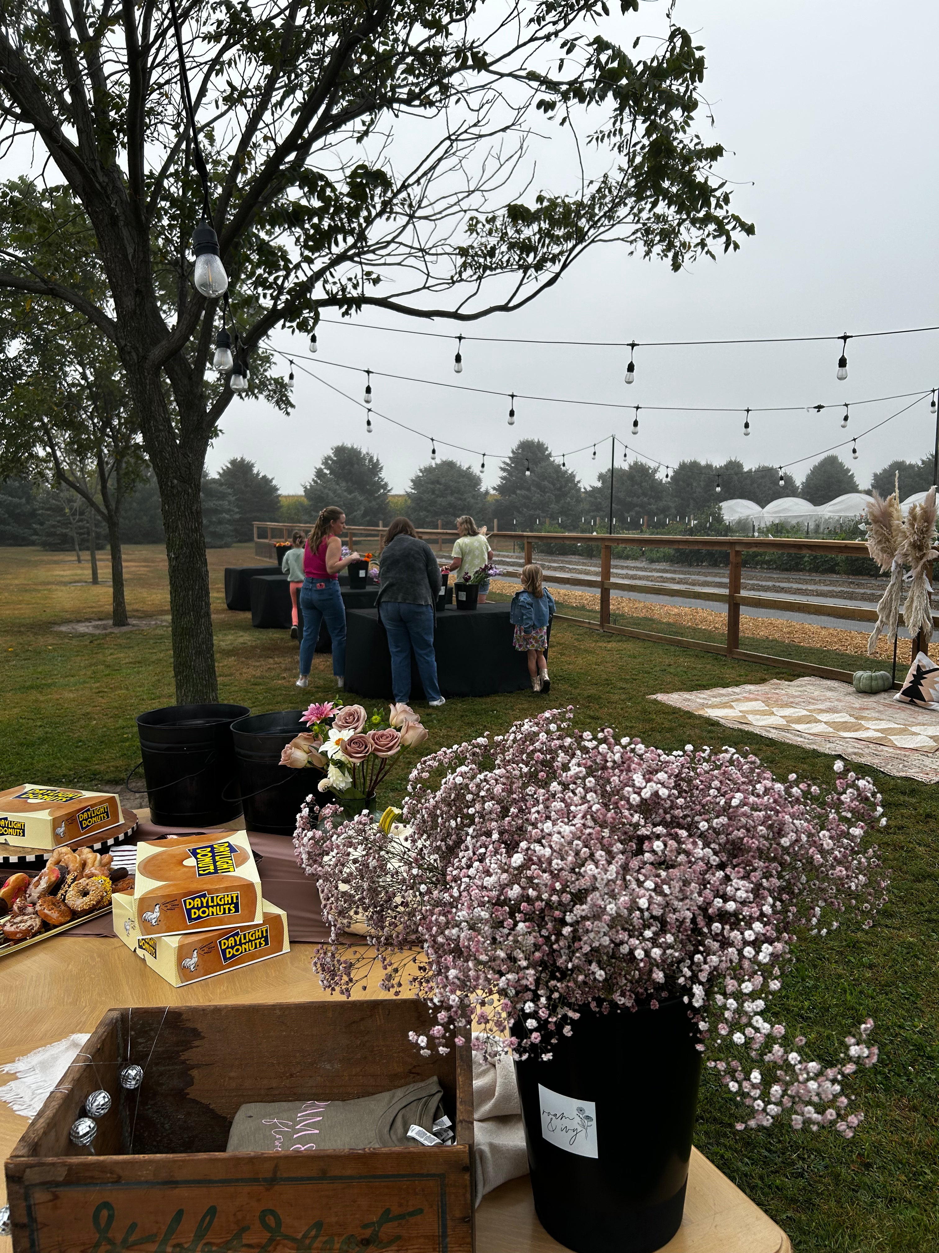 Outdoor market scene with tables, flowers, people browsing, and string lights hanging from trees.