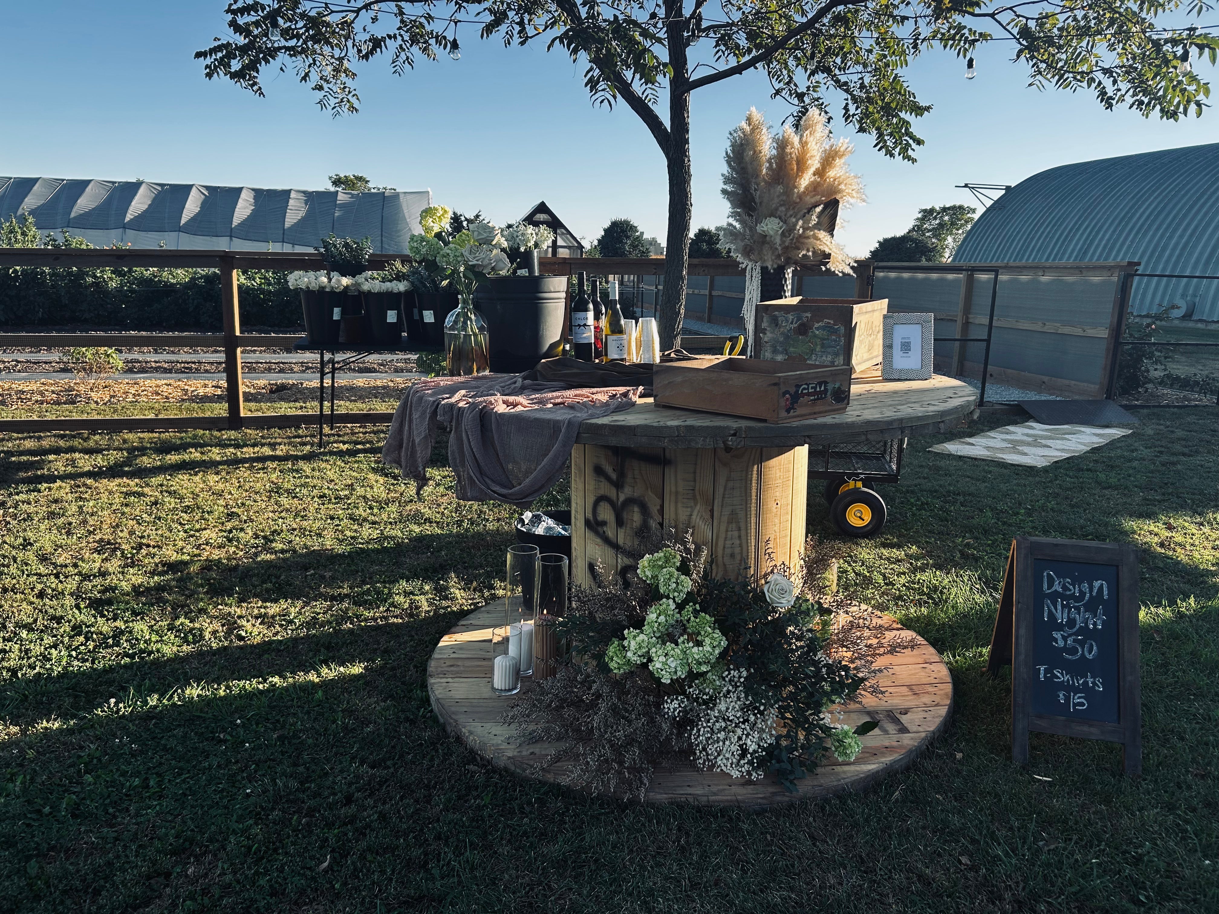 Outdoor wooden table display with flowers and candles, set up in a grassy area. Nearby is a chalkboard sign listing prices for "Design Night" and T-shirts. Barns are visible in the background.