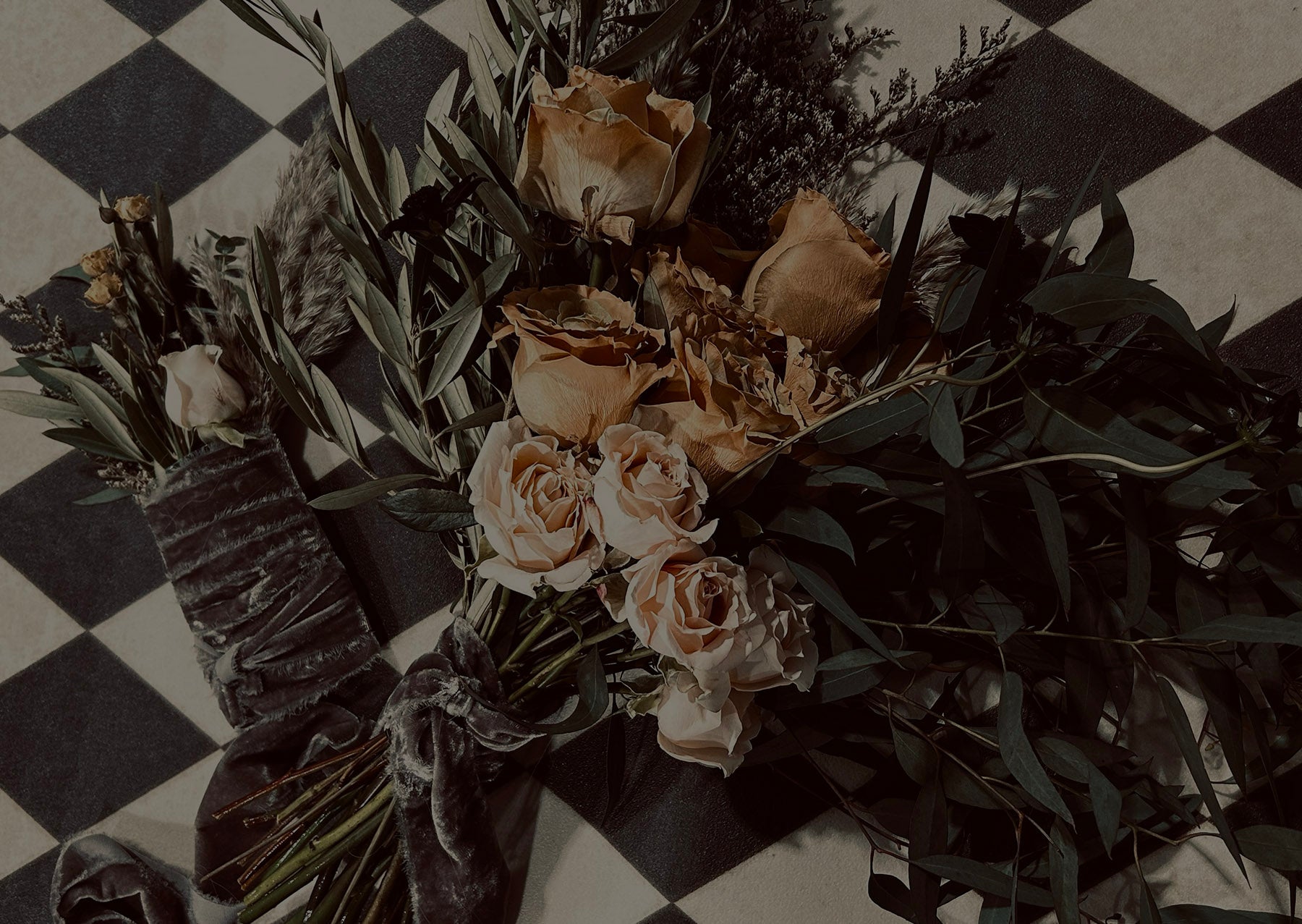 A bouquet of faded roses with greenery and a velvet ribbon rests on a black and white checkered surface.